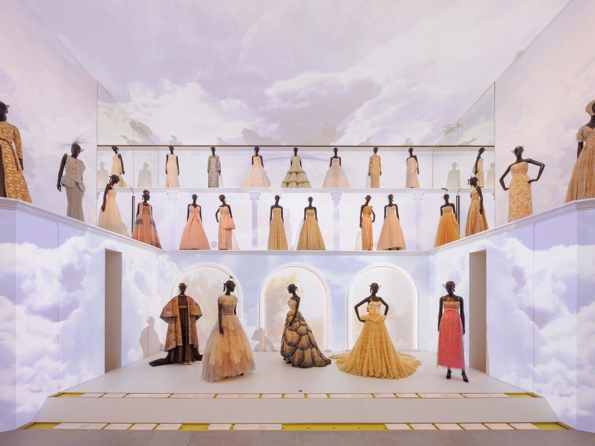 Mannequins in couture gowns displayed on tiered platforms in a white gallery painted with clouds
